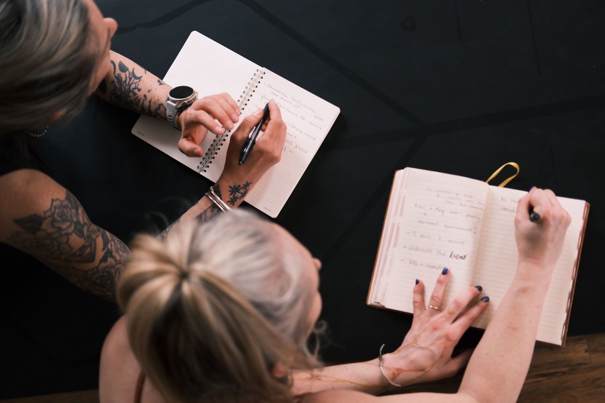 Two yoga teachers writing class plans by hand in their notebooks on a black mat, seen from above